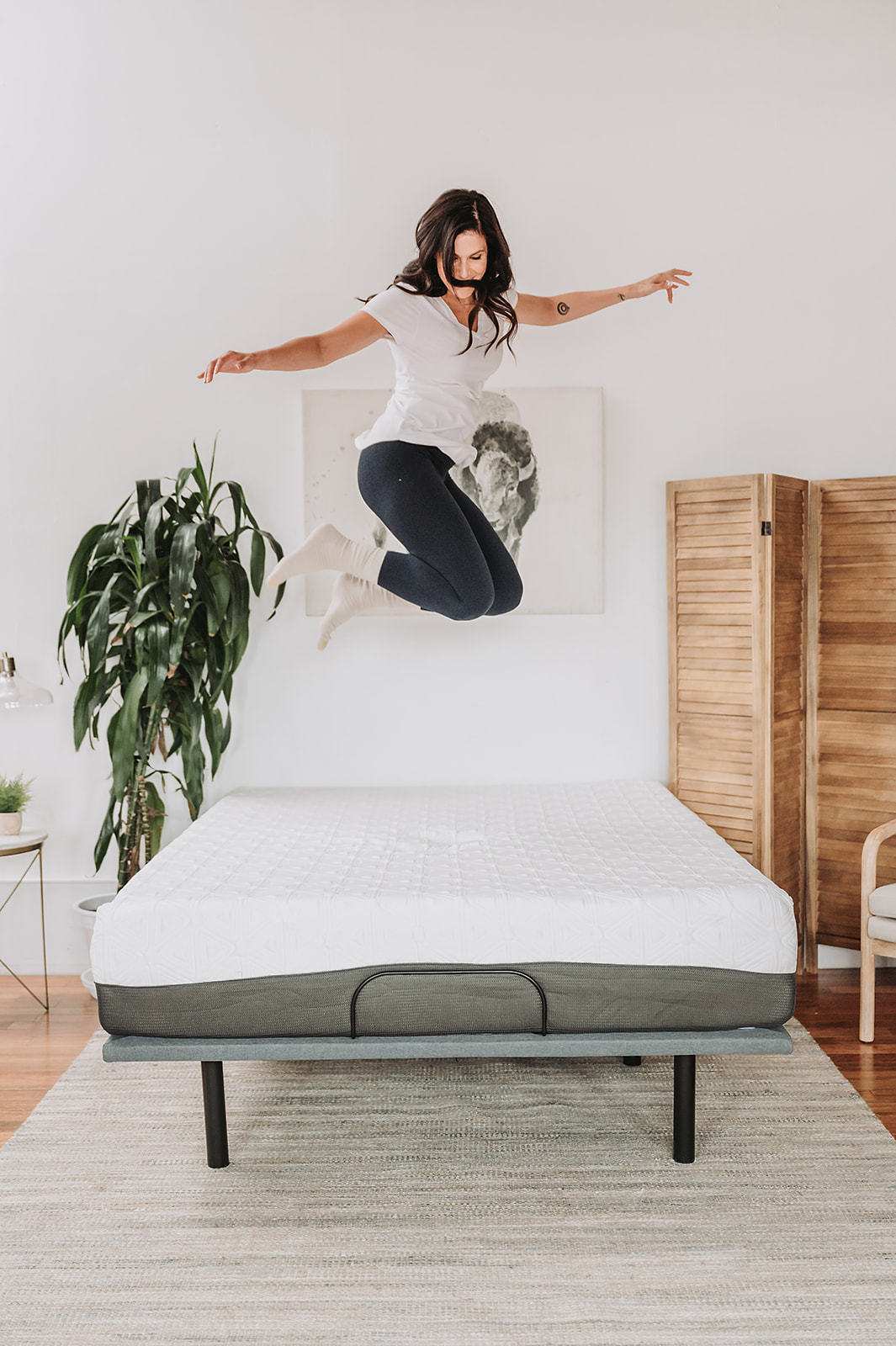 Woman jumping on a modern mattress in a bright bedroom with house plants and wood decor